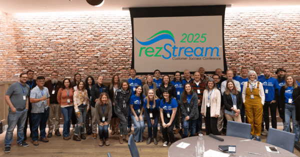 Group photo of attendees and staff at the 2025 rezStream Customer Success Conference, smiling together in front of a presentation screen displaying the event logo at the Maven Hotel in Denver.
