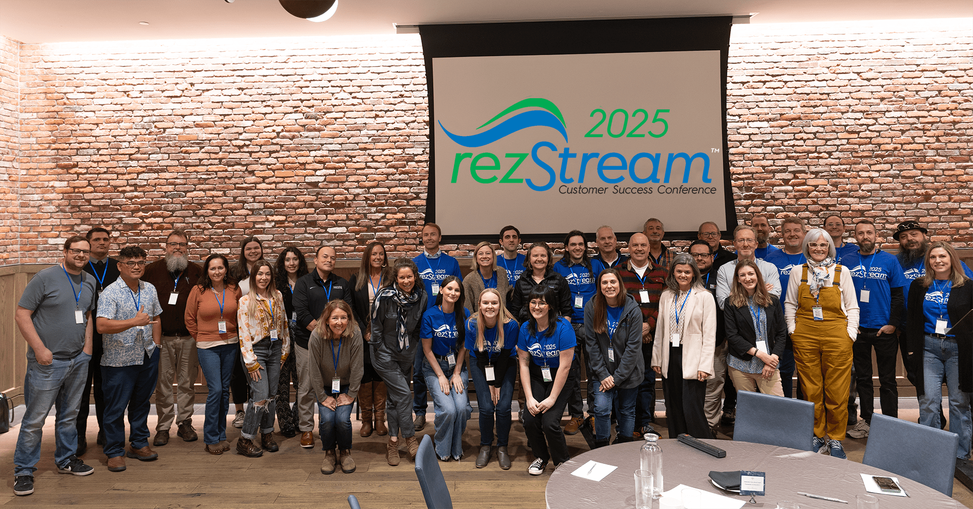 Group photo of attendees and staff at the 2025 rezStream Customer Success Conference, smiling together in front of a presentation screen displaying the event logo at the Maven Hotel in Denver.